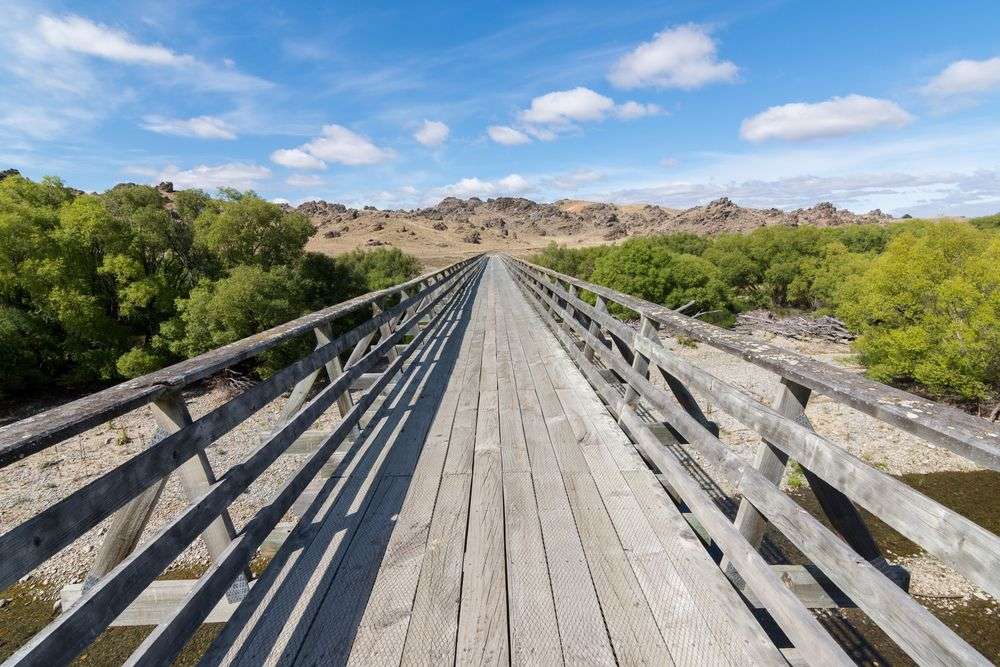 Eine Holzbrücke über einen Fluss mit Bäumen und Bergen im Hintergrund