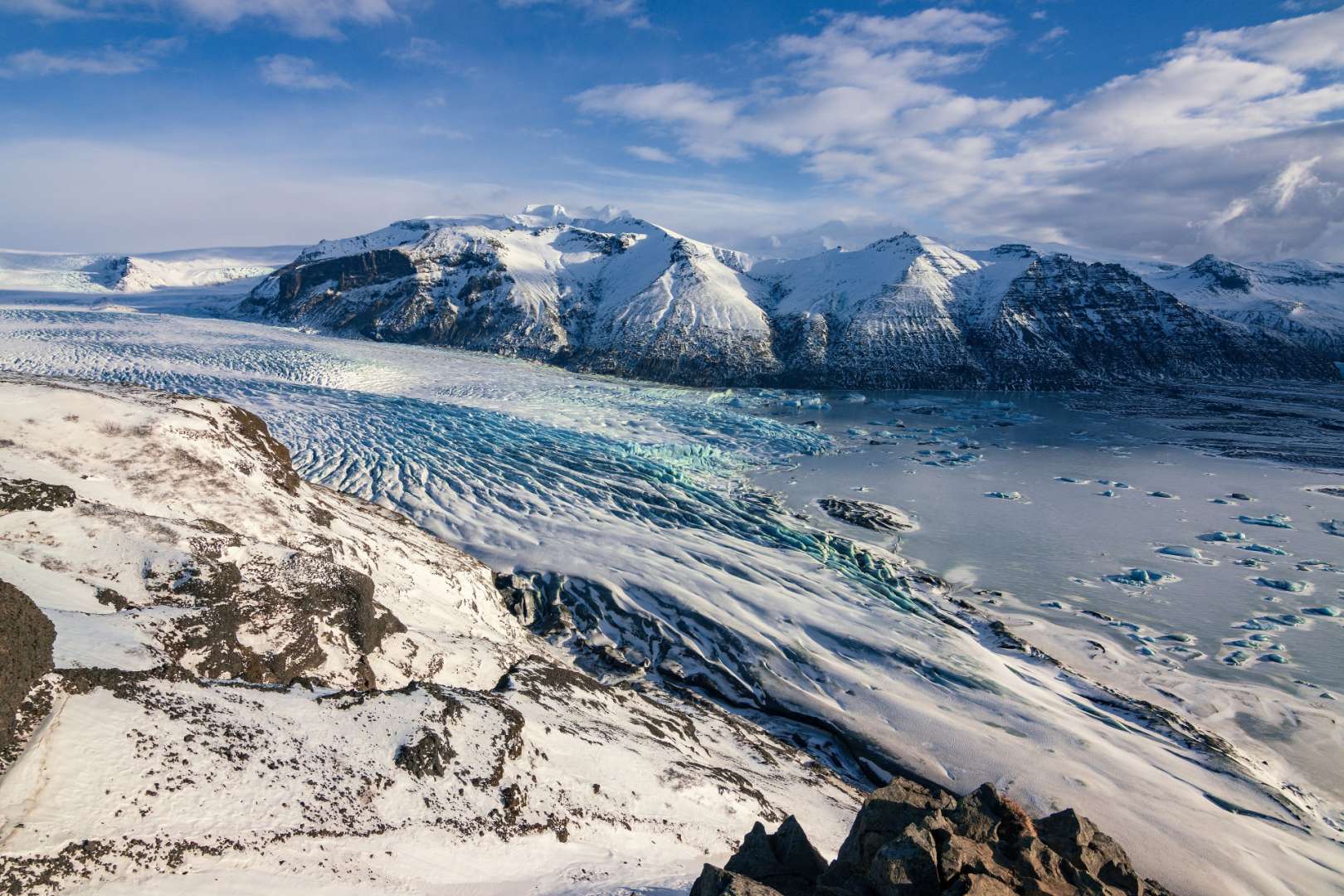 Eine weite Gletscherzunge erstreckt sich in eine eisbedeckte Berglandschaft unter einem blauen Himmel mit Wolken.