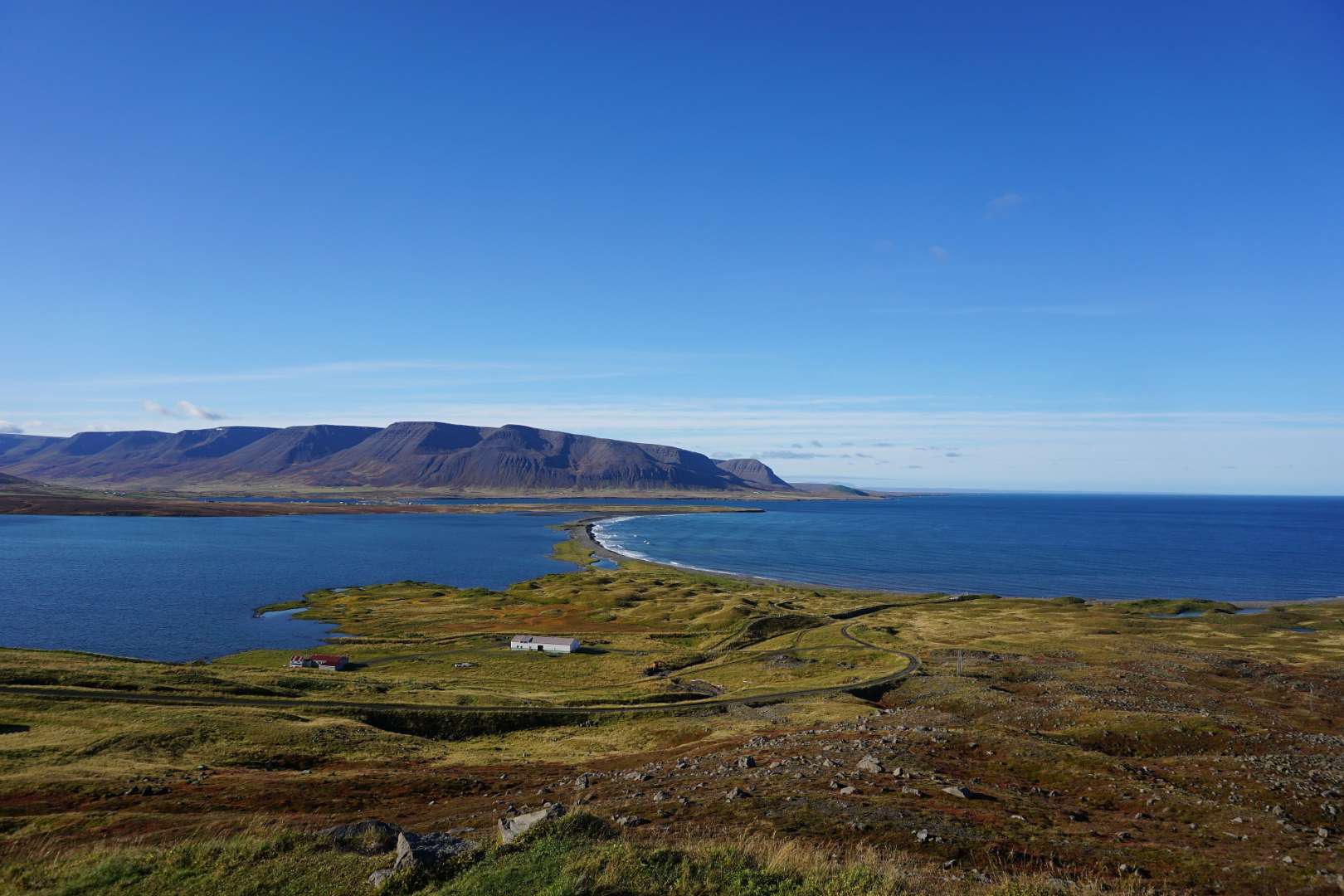 Ein weites Panorama zeigt eine Küstenlandschaft mit blauer See, grünen Hügeln, einer schmalen Landzunge, Bergen im Hintergrund und einem strahlend blauen Himmel.