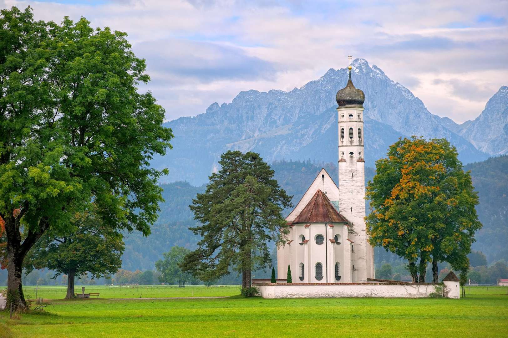 Eine kleine weiße Kirche steht inmitten eines grünen Feldes mit Bäumen und Bergen im Hintergrund