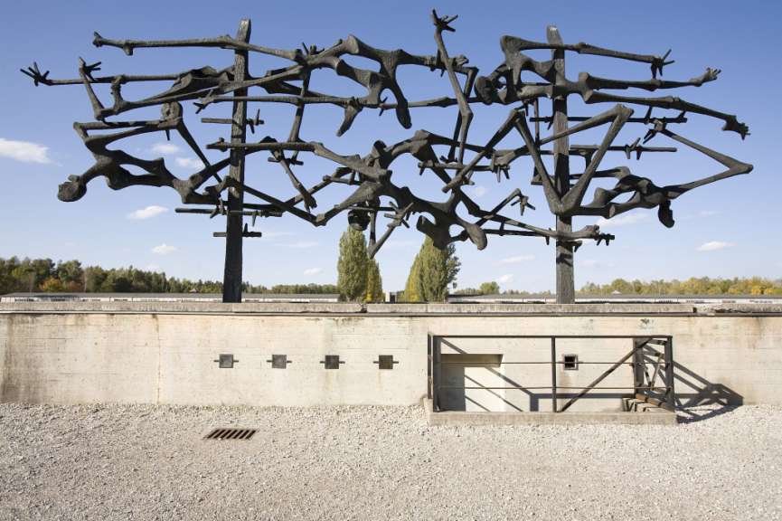 Eine dunkle, abstrakte Skulptur, die gefolterte Figuren darstellt, steht vor einer Betonmauer und einer Reihe von Gebäuden im Hintergrund unter einem blauen Himmel.