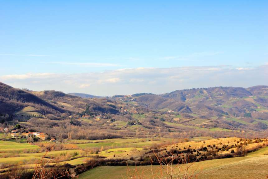 Eine weite Hügellandschaft mit spärlich bewachsenen Bäumen, Feldern und kleinen Dörfern unter einem blauen Himmel mit dünnen Wolken.