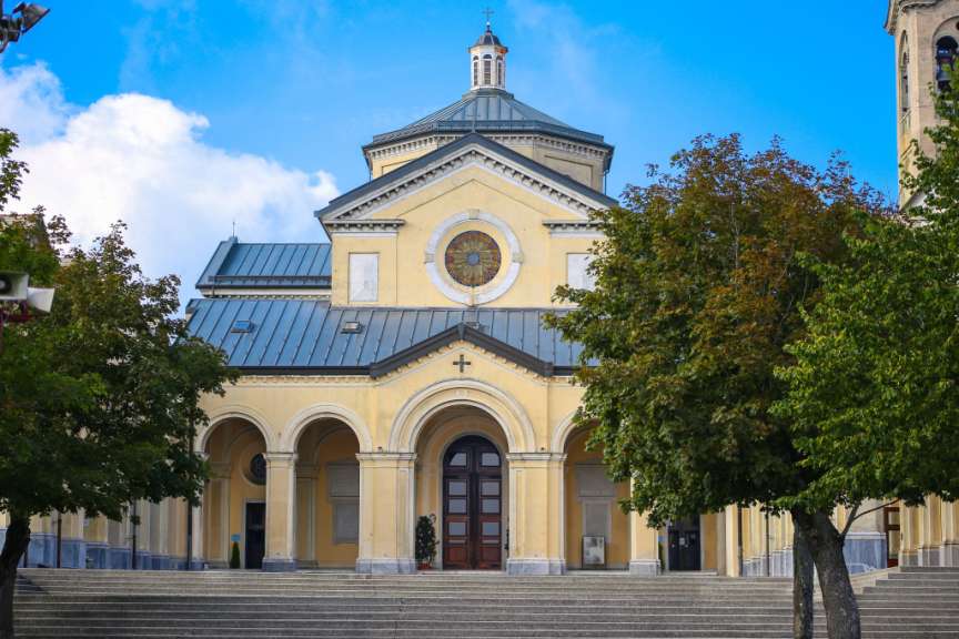 Eine gelbe Kirche mit einer Kuppel und einem Glockenturm wird von Bäumen und blauen, bewölkten Himmel umgeben.