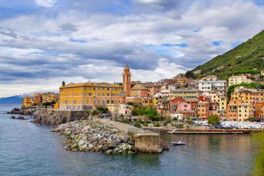 Ein italienisches Küstendorf mit bunten Häusern, das sich an einen grünen Hügel schmiegt, mit einem kleinen Hafen voller Boote und einem steinigen Ufer.