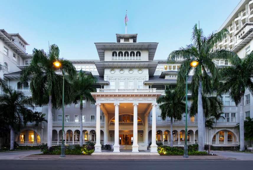 Dieses Bild zeigt den prächtigen Eingangsbereich des Moana Surfrider, A Westin Resort & Spa, Waikiki Beach, mit seinen eleganten Säulen, Balustraden und umliegenden Palmen im Abendlicht.