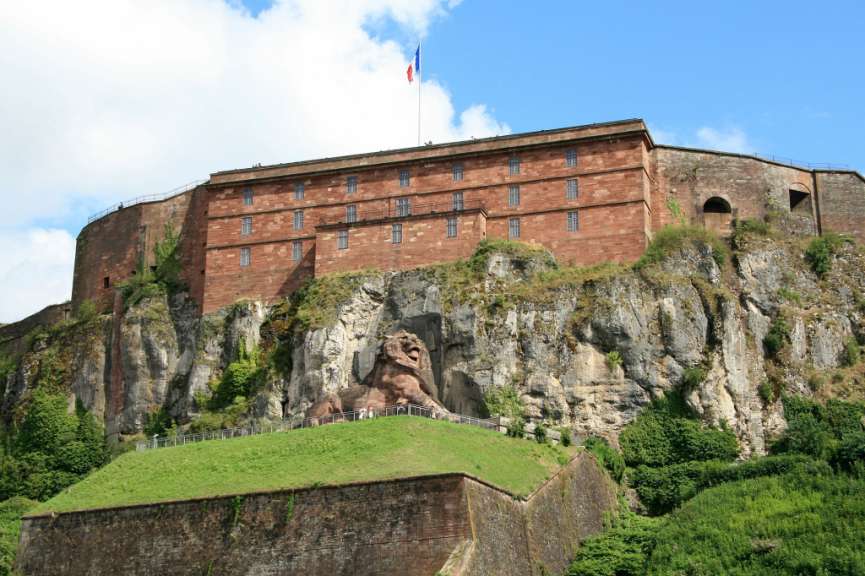 Eine Festung mit einem steinernen Löwenrelief, der auf einer Felswand thront, wird von unten nach oben gezeigt, mit einem blauen und roten französischen Fahnen im Hintergrund.
