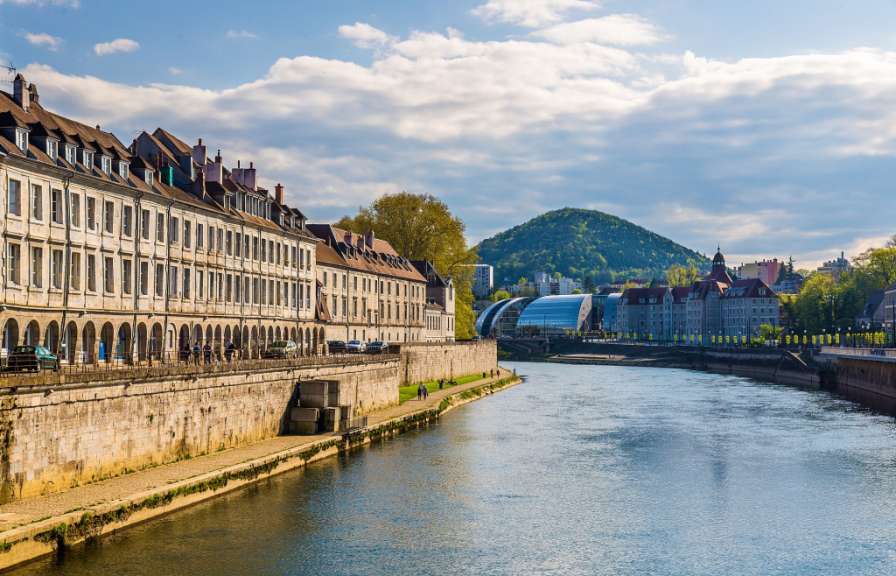 Eine breite Flussuferpromenade mit einem Gebäude auf der linken Seite und einer begrünten Hügelkuppe im Hintergrund.