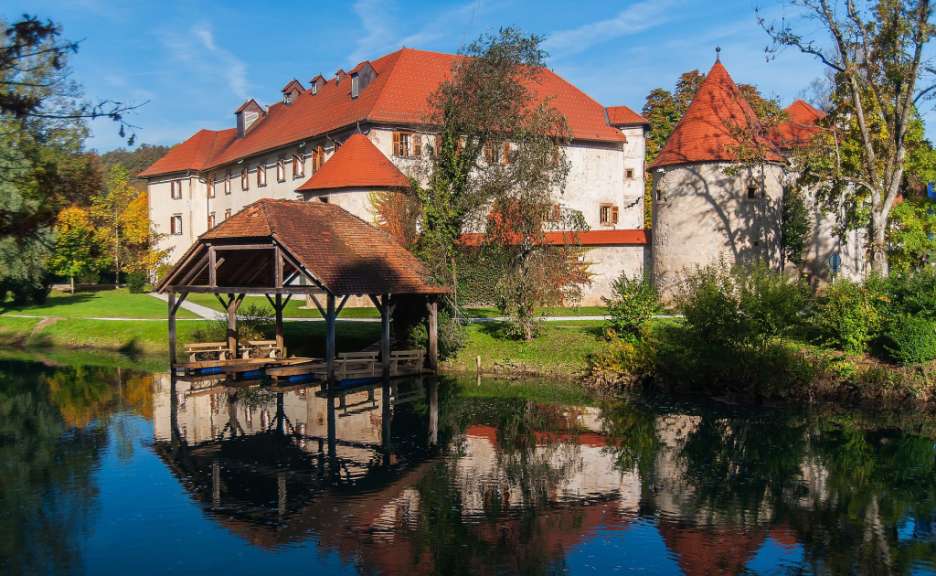 Ein Schloss mit roten Dächern spiegelt sich im ruhigen Wasser eines Sees, vor dem ein kleiner Bootssteg mit einem Dach steht.