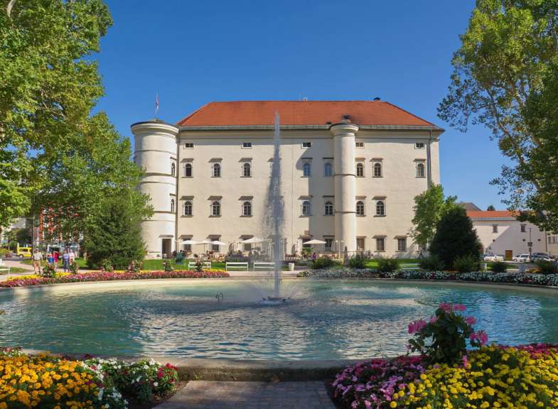 Ein großer Wassergarten mit einem Brunnen und einem Schloss im Hintergrund wird von Blumen und Bäumen eingerahmt.