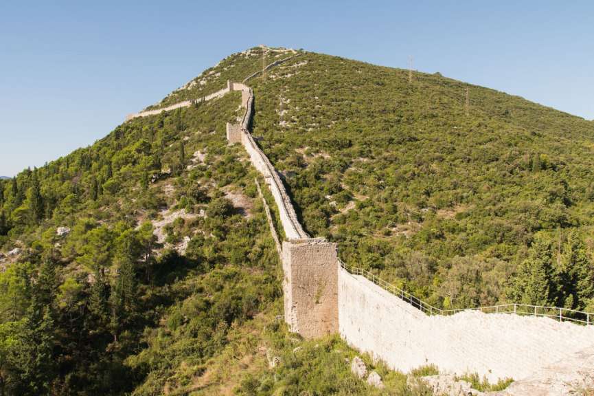 Eine lange, steinerne Mauer schlängelt sich einen mit grüner Vegetation bewachsenen Hügel hinauf unter einem klaren blauen Himmel.