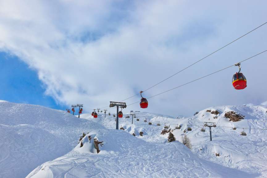 Eine Reihe von roten Gondeln fährt auf einem Skigebiet an schneebedeckten Berghängen unter einem bewölkten blauen Himmel.
