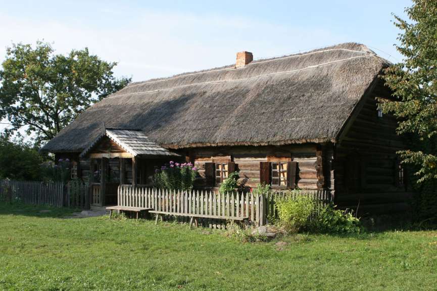Ein traditionelles Holzhaus mit Reetdach und Holzzaun steht auf einer grünen Wiese vor einem blauen Himmel.