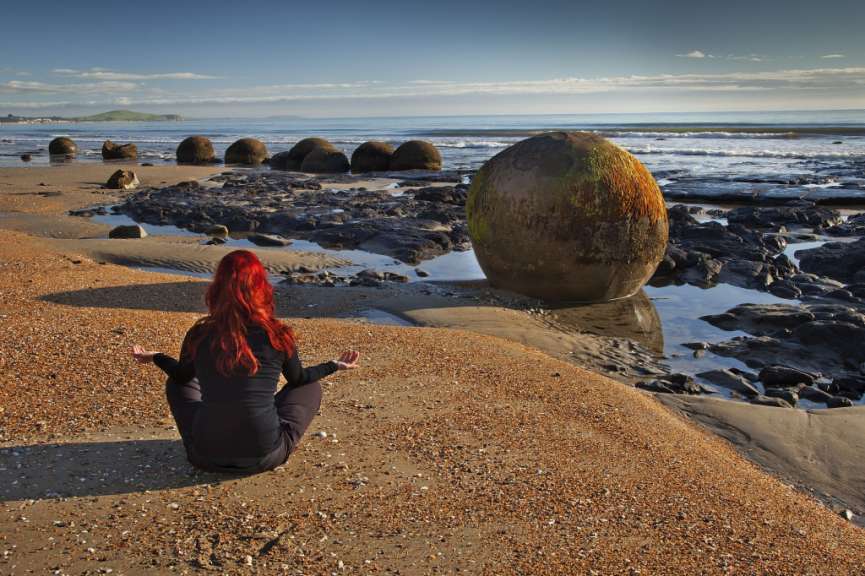 Eine Frau mit roten Haaren meditiert auf einem Strand in der Nähe von großen, runden Felsbrocken am Meer.