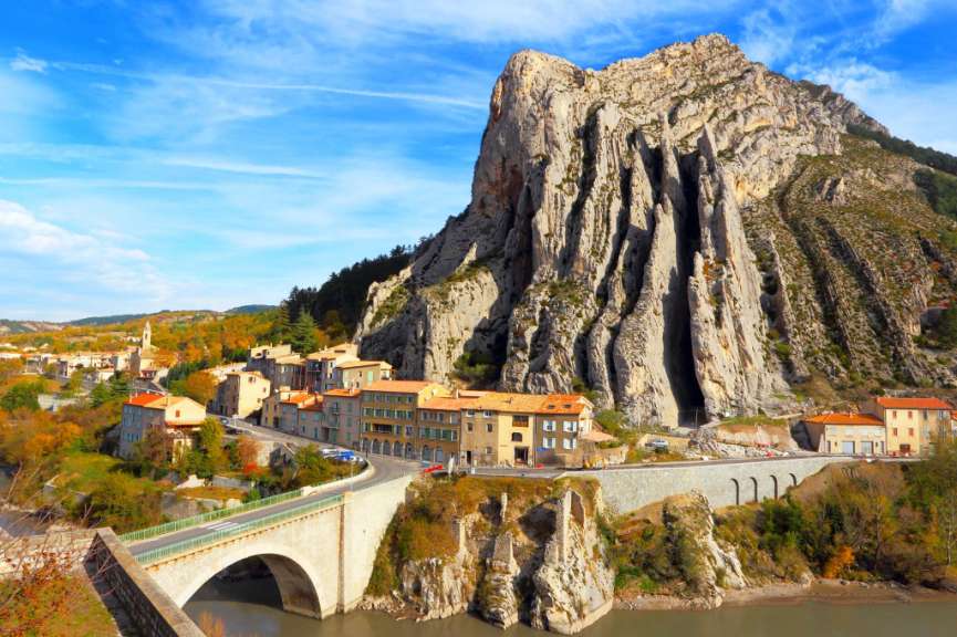 Eine malerische französische Stadt schmiegt sich an einen majestätischen Felsen, überragt von einem strahlend blauen Himmel mit vereinzelten Wolken, während sich ein Fluss friedlich durch die Landschaft schlängelt.