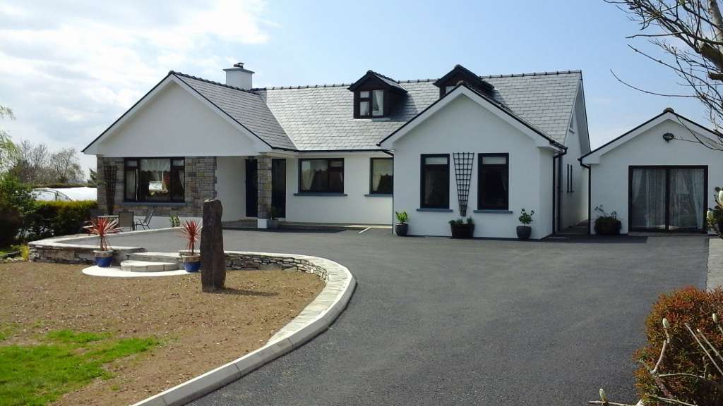 Ein weißes Einfamilienhaus mit dunklem Schieferdach und einem großen, asphaltierten Vorplatz steht an einem sonnigen Tag vor einer grünen, hügeligen Landschaft.
