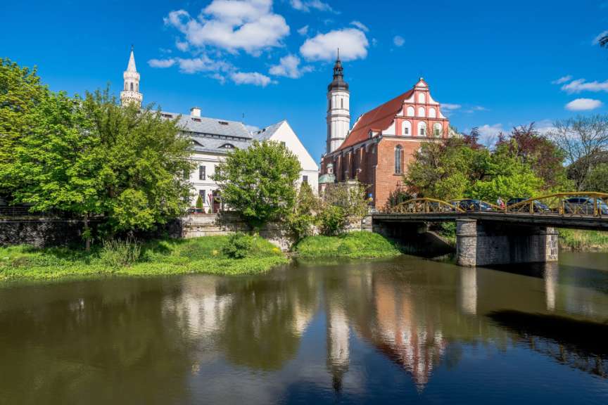 Zwei Kirchen mit Türmen, eine Brücke und Bäume spiegeln sich in einem Fluss vor blauem Himmel.