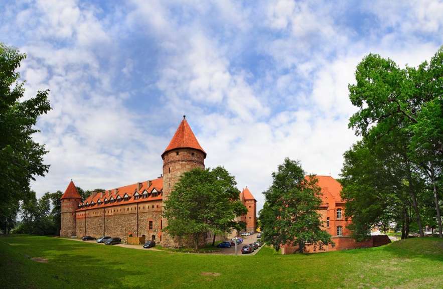 Eine weite Aufnahme zeigt ein steinernes Schloss mit roten Ziegeldächern und Türmen, umgeben von grünen Bäumen und einer weitläufigen Rasenfläche.