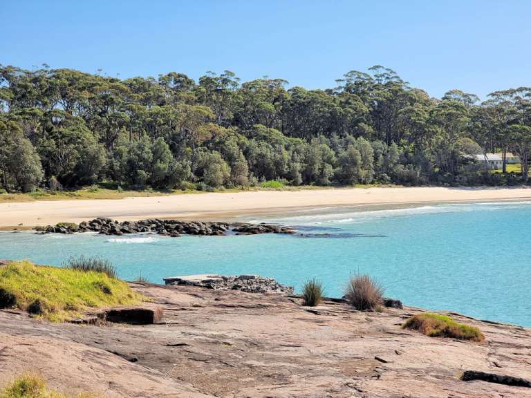 Ein sonniger Tag mit einer ruhigen Küstenlandschaft, bestehend aus einem Sandstrand, einem türkisfarbenen Meer und einem dichten Wald im Hintergrund.