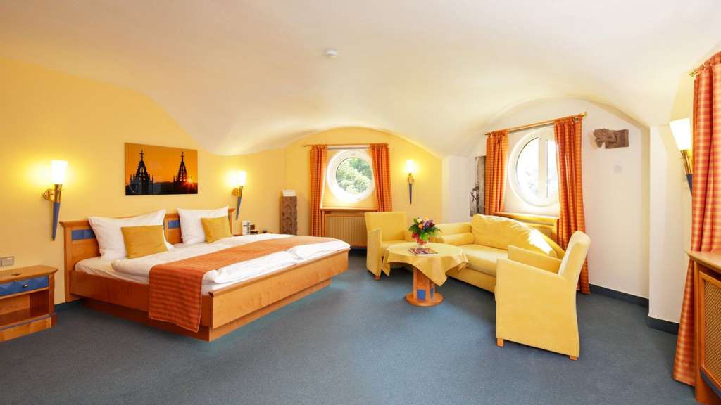 Eine geräumige Suite mit einem großen Bett, einer gemütlichen Sitzecke mit Sofa und Sesseln sowie runden Fenstern, dekoriert mit orangen Vorhängen und einem Gemälde von Kirchtürmen.