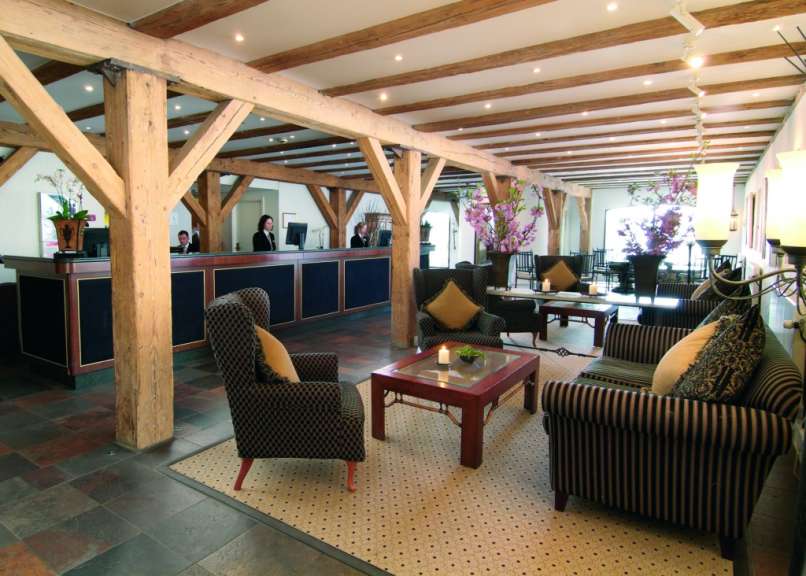 In einer Hotellobby mit Holzbalken und einem Empfangsbereich sitzen Gäste in bequemen Sesseln.