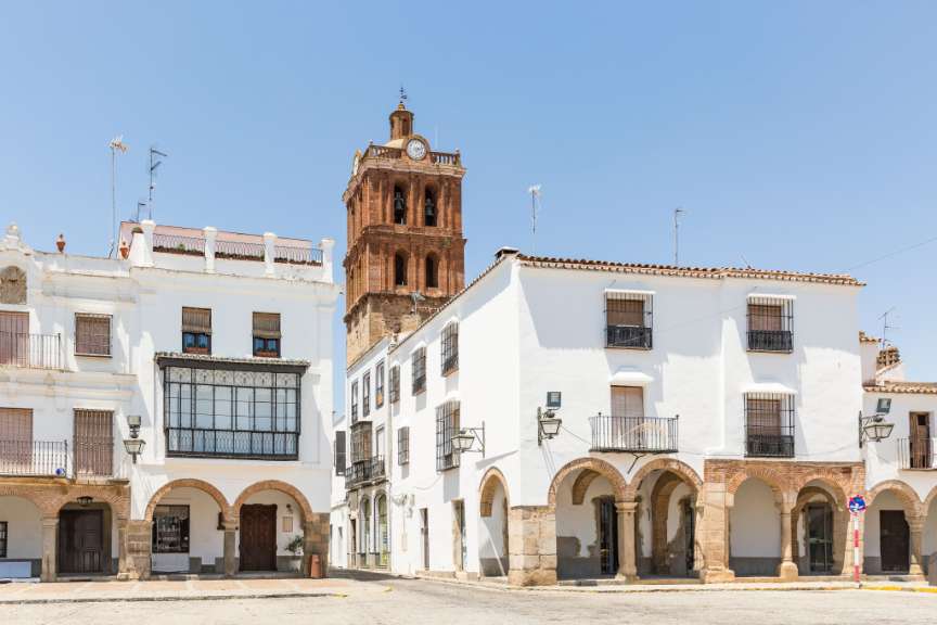 In einer spanischen Stadt mit weißen Gebäuden und Arkaden ist ein Kirchturm aus rotem Ziegelstein zu sehen.