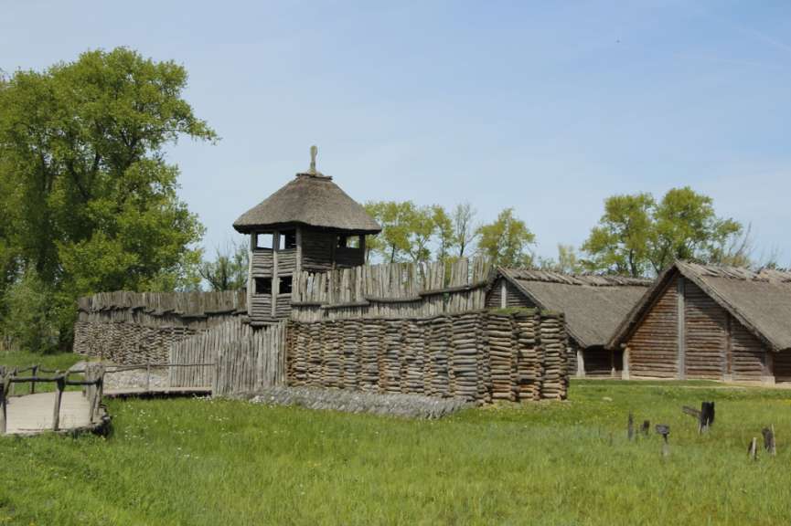 Eine hölzerne Festung mit einem Wachturm und zwei strohgedeckten Häusern steht auf einer grünen Wiese unter blauem Himmel.