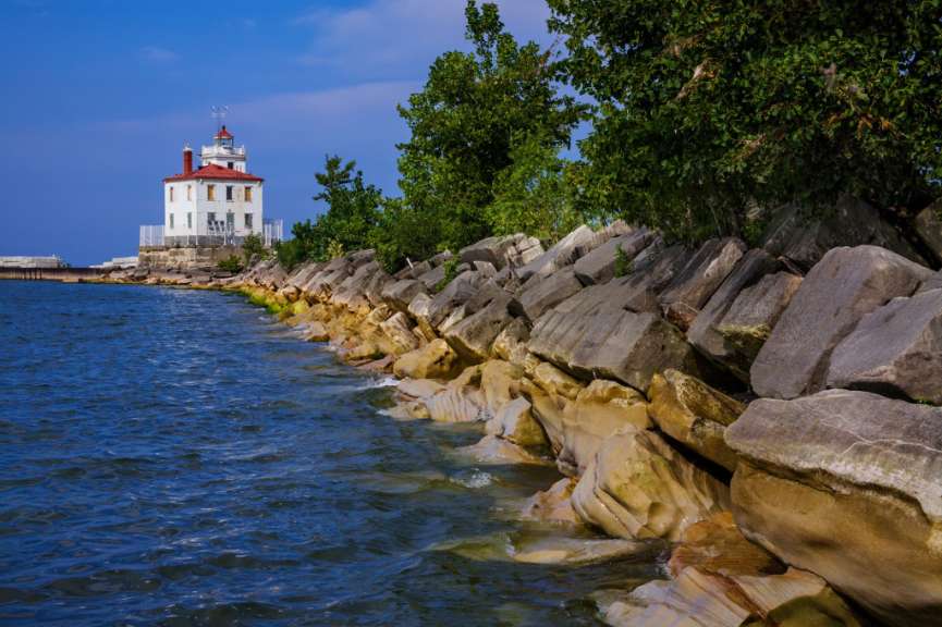 Ein weißer Leuchtturm mit rotem Dach steht auf einer felsigen Halbinsel am Ufer eines blauen Sees.