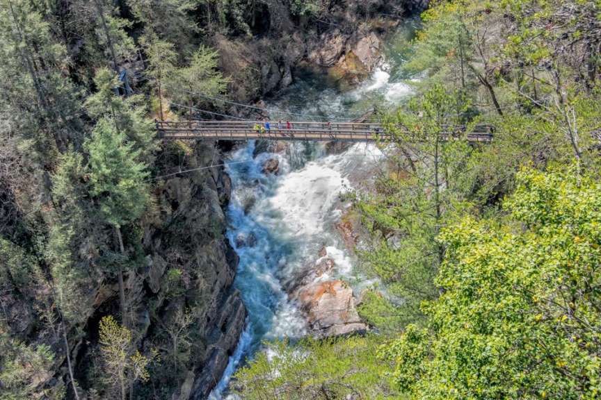 Ein Fluss fließt durch eine Schlucht mit Felsen und Bäumen, über den eine Fußgängerbrücke führt, auf der einige Personen spazieren gehen.