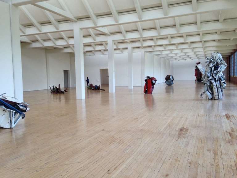 In einer großen Ausstellungshalle stehen mehrere abstrakte Skulpturen auf einem Holzfußboden.