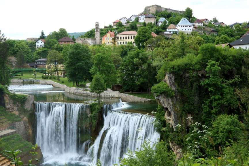 Ein malerisches Dorf mit traditionellen Häusern und einer Burgruine thront über einem breiten Wasserfall im türkisfarbenen Wasser.
