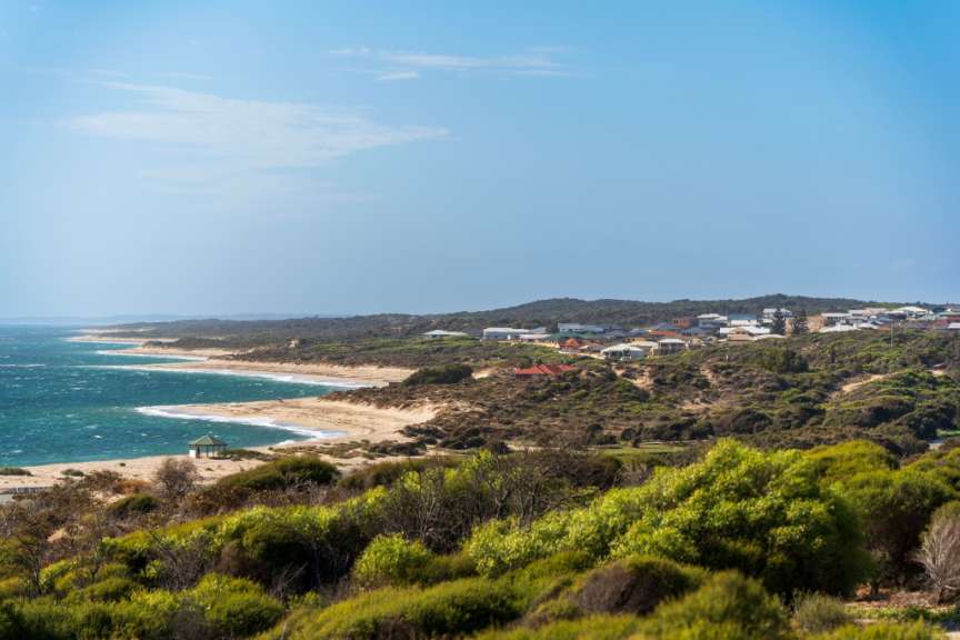 Ein Blick über bewaldete Hügel und Sanddünen auf eine Bucht mit türkisem Wasser, an der sich eine Wohnsiedlung und ein kleiner Pavillon befinden.