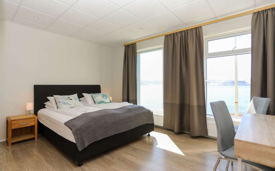 Ein Schlafzimmer mit großem Bett, Nachttisch und Blick durch die Fenster auf das Meer und die Küste.