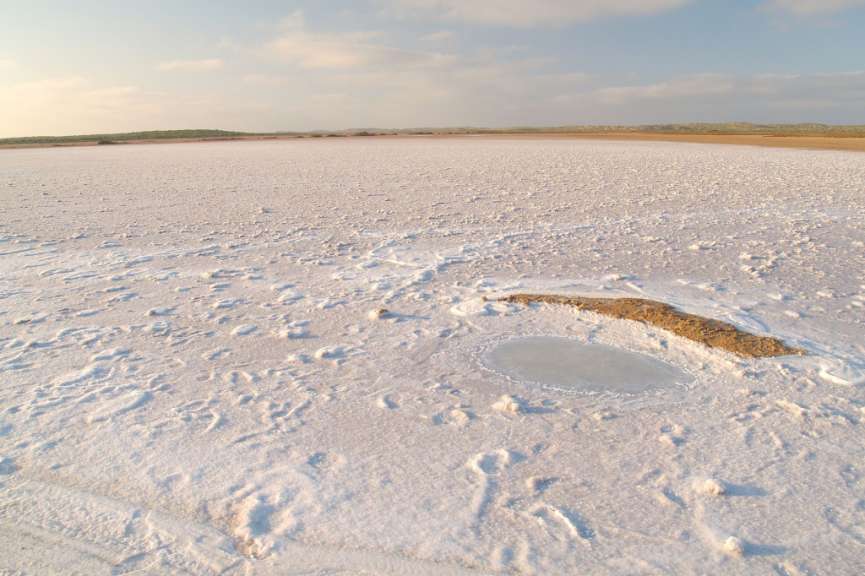 Eine weite Salzfläche mit vereinzelten kleinen Wasserlöchern und einer aufgeschütteten Erdformation erstreckt sich unter einem wolkenverhangenen Himmel.