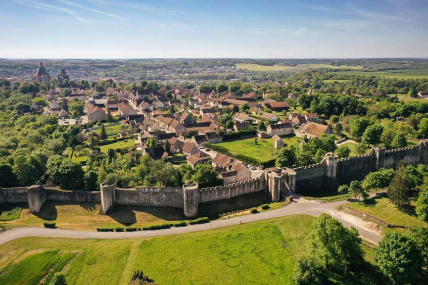 Eine Drohnenaufnahme zeigt eine mittelalterliche Stadtmauer, die eine Stadt mit traditionellen Häusern und grünen Bäumen umschließt, vor einer sanften Hügellandschaft.