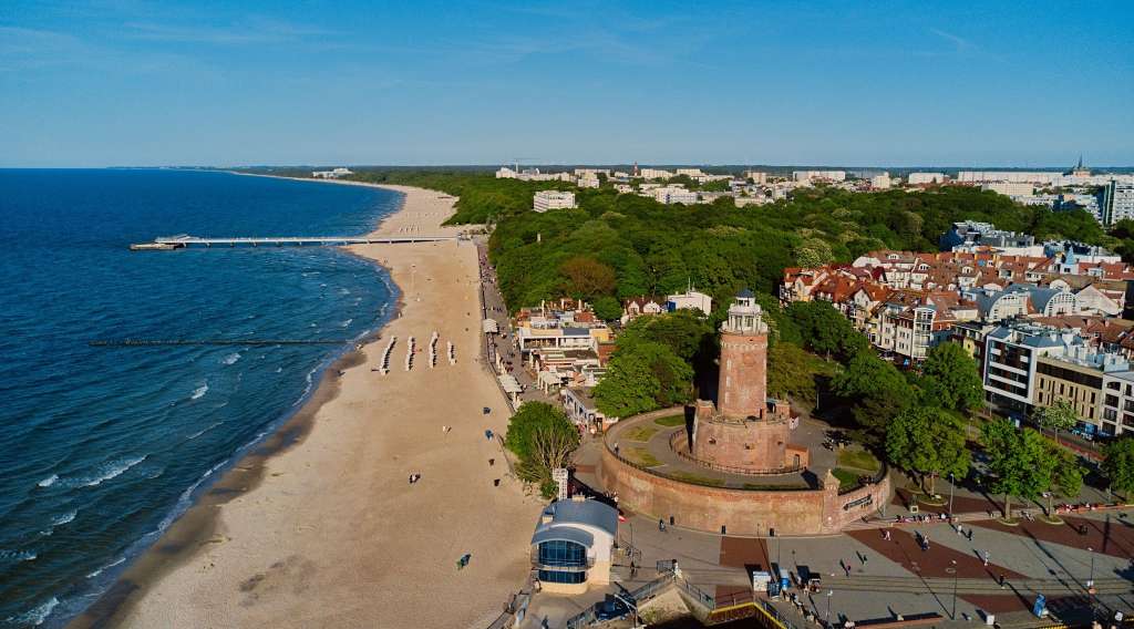 Eine Drohnenaufnahme zeigt einen breiten Sandstrand mit dem Meer, einen Pier, eine Stadt mit Bäumen und einen charakteristischen Leuchtturm.