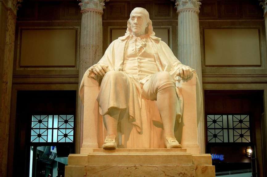 Eine weiße Marmorstatue von Benjamin Franklin sitzt in einem großen Gebäude mit Säulen.