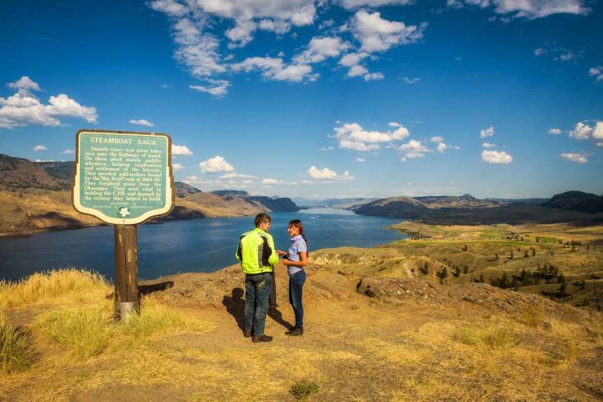 Ein Mann und eine Frau stehen auf einem Aussichtspunkt und blicken über einen See und eine bergige Landschaft, während sie eine Informationsstele betrachten.
