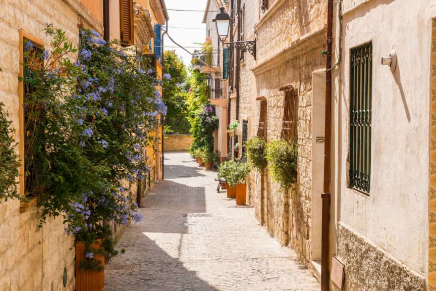 Ein schmaler, gepflasterter Weg führt durch eine Gasse, die auf beiden Seiten von traditionellen Steinhäusern gesäumt wird, und auf einer Seite wuchert eine üppige blaue Blumenranke.