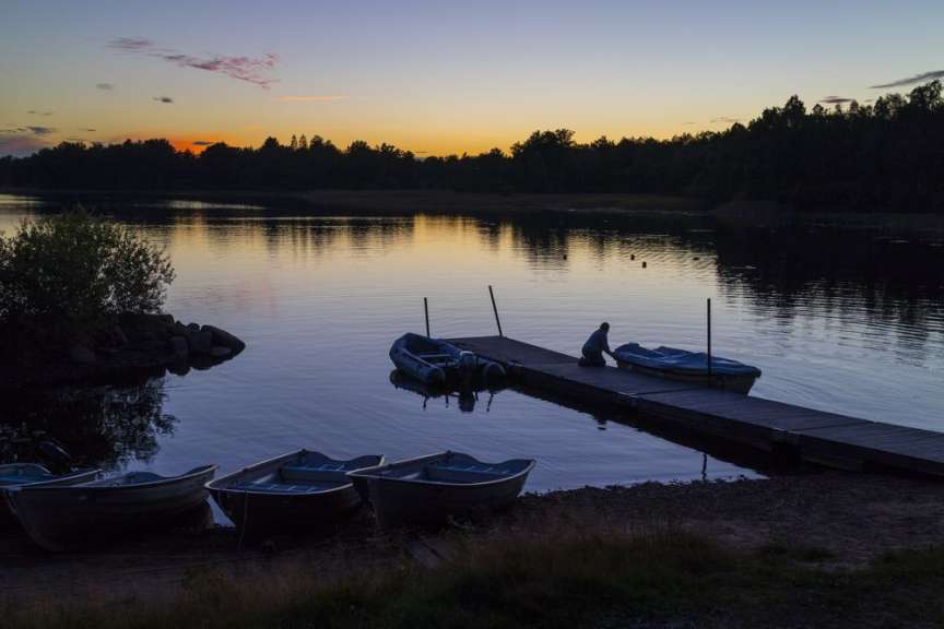 Eine Person kniet auf einem Steg und bedeckt ein Boot mit einer Plane, während hinter ihr ein ruhiger See und ein Dämmerungshimmel zu sehen sind.