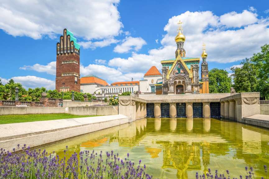 Eine Ansicht des Künstlerkolonie Mathildenhöhe in Darmstadt mit einem turmartigen Gebäude, einer Kapelle mit goldenen Kuppeln und einem reflektierenden Teich im Vordergrund.