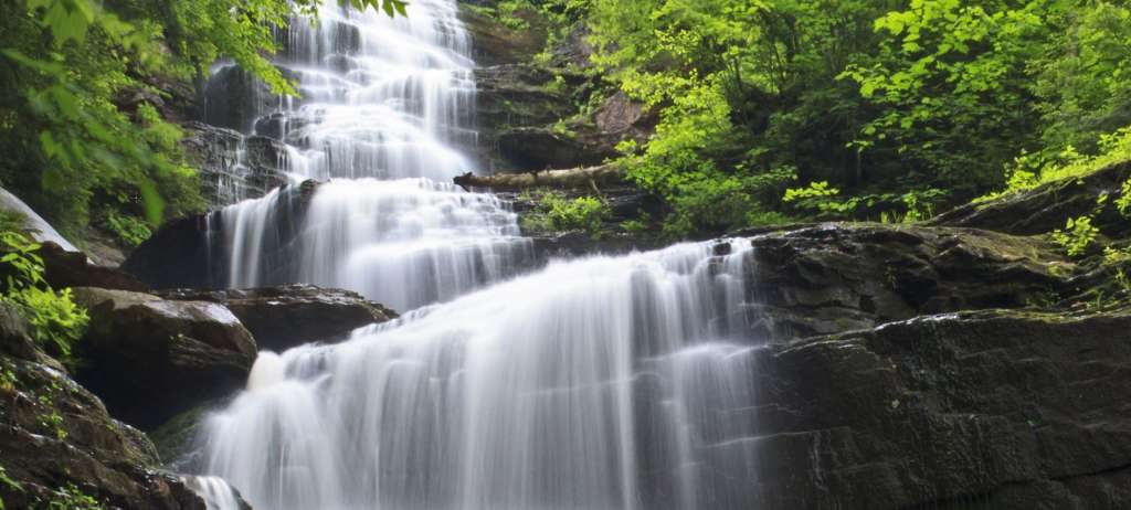Ein breiter Wasserfall stürzt in mehreren Stufen über dunkle Felsen in üppige, grüne Vegetation.