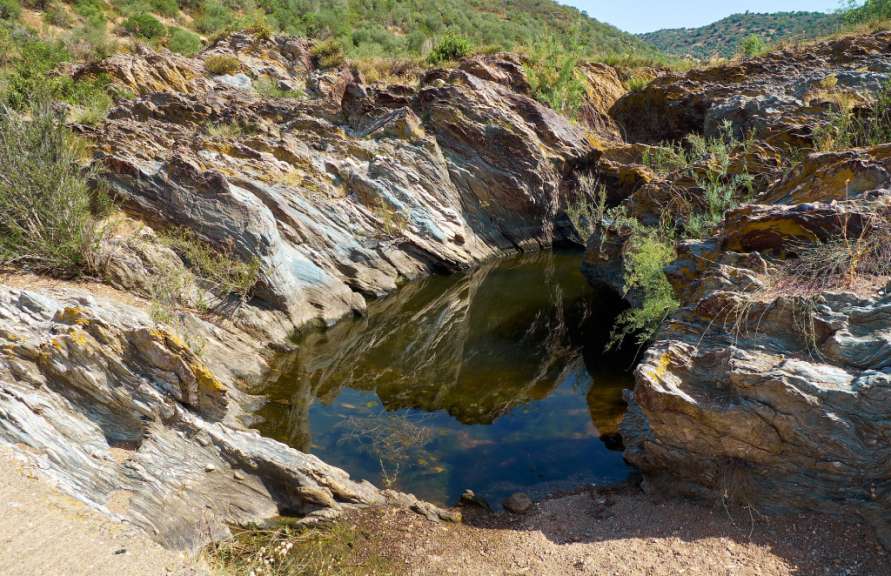 Ein kleiner Teich spiegelt sich in schattigen Felsen, während am Hang darüber grüne Vegetation wächst.