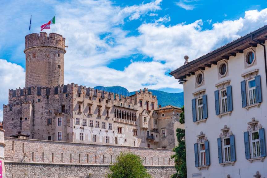 Auf diesem Bild ist eine Burg mit einem runden Turm und einer daran angebauten Stadtmauer zu sehen, während im Vordergrund ein helles Gebäude mit blauen Fensterläden steht.