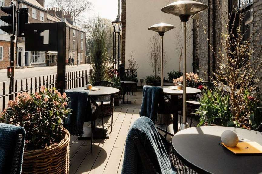 Auf einer sonnigen Terrasse stehen runde Tische mit Sitzgelegenheiten, Heizstrahlern und Pflanzen vor einer Straße mit Gebäuden.