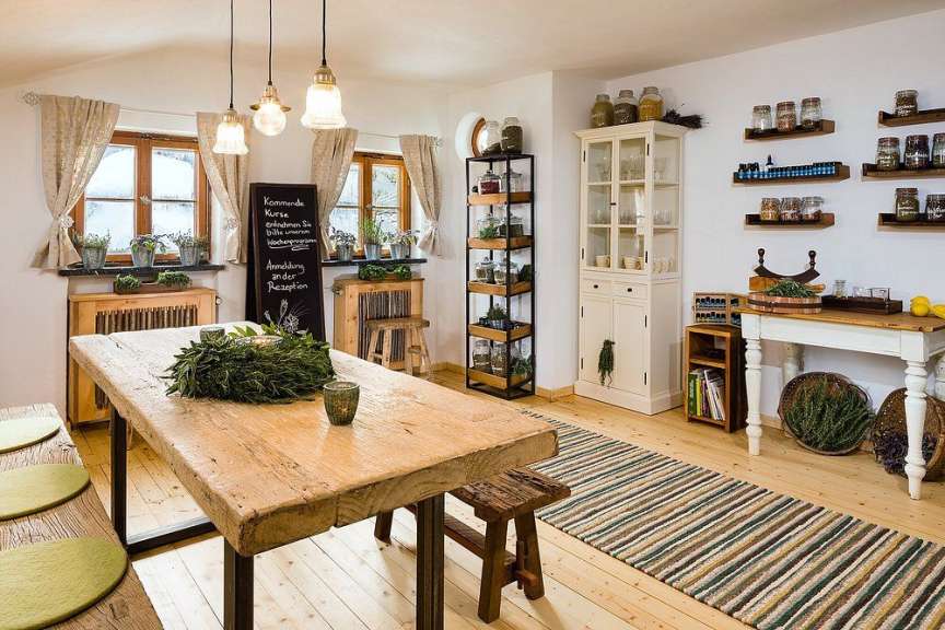 In einem rustikalen Raum steht ein großer Holztisch, dekoriert mit einem Kranz aus Kräutern, und ein Regal mit Gläsern sowie ein Schrank sind im Hintergrund zu sehen.