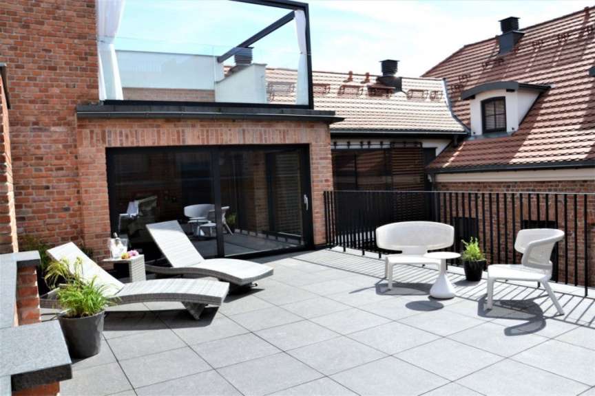 Auf einer Dachterrasse mit Ziegelsteinmauer und modernen Möbeln stehen zwei Sonnenliegen, ein Tisch mit einer Flasche Champagner und zwei weiße Stühle.