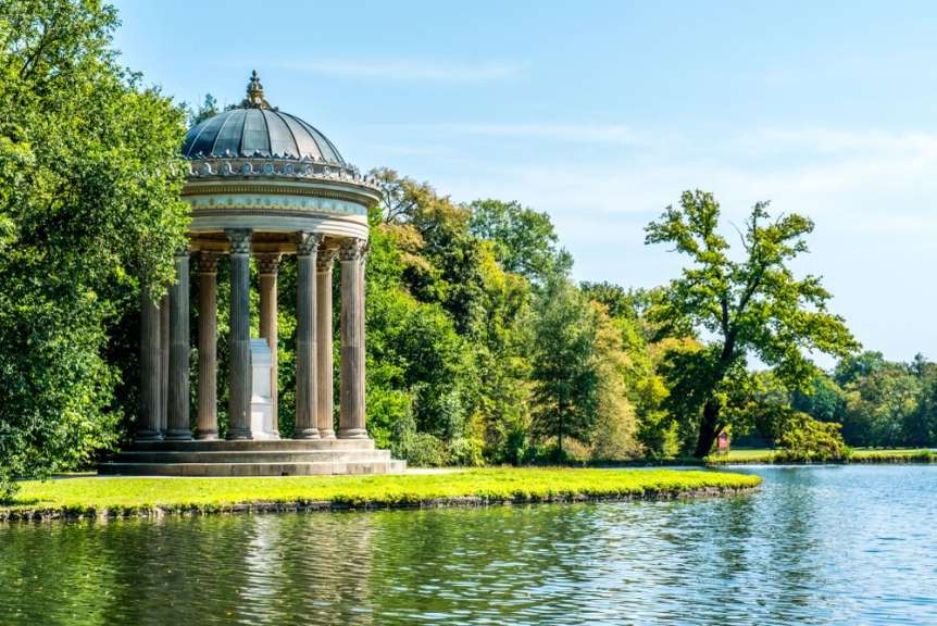 Ein antiker Monopteros steht am Ufer eines ruhigen Sees, umgeben von üppigem Grün und unter einem klaren blauen Himmel.