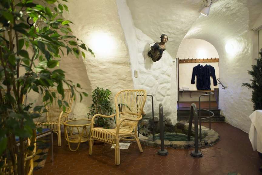 In einem Ruheraum mit rustikalen Steinwänden und gewölbter Decke stehen Korbstühle, ein kleiner Tisch und ein Becken mit Wasserspielen, daneben ein Treppenaufgang mit Bademänteln.