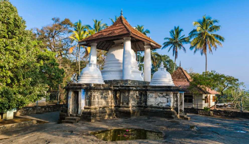 Ein buddhistischer Stupa mit mehreren kleineren Stupas und einem Dach steht auf einem Felsen vor Palmen und Bäumen.