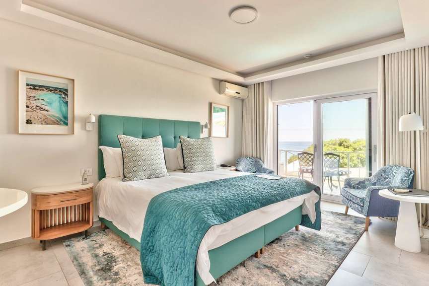 Ein großes Bett mit türkisfarbener Tagesdecke und Kissen steht in einem hellen Schlafzimmer mit Blick auf das Meer.